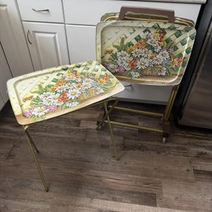 May include: A set of rectangular metal TV tray tables with gold-toned legs and a floral pattern featuring white daisies, yellow flowers, and green foliage against a white lattice background. One tray is standing upright in a storage rack.