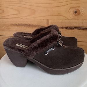 Pode incluir: Tamancos Skechers castanhos com acabamento em pele sintética. Os tamancos têm bico arredondado, salto bloco e detalhe de tira com ilhós prateados. O logótipo Skechers é visível na lateral do sapato.