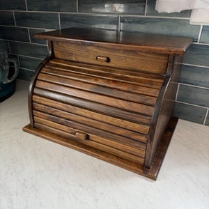 May include: A dark brown wooden bread box featuring a roll-top door with horizontal slats and a small handle. The rectangular storage container has a flat top surface and a solid base, showcasing a natural wood grain finish throughout the structure.