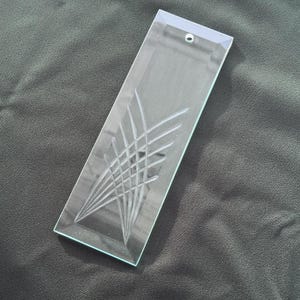 May include: A clear, rectangular glass ornament with an etched geometric design. The ornament has a small hole at the top for hanging. The etched design features a series of intersecting lines, creating a modern, abstract pattern.