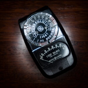 May include: A black and silver General Electric light meter with a dial that reads "Light Values" and "Cover Closed" to "Cover Open".