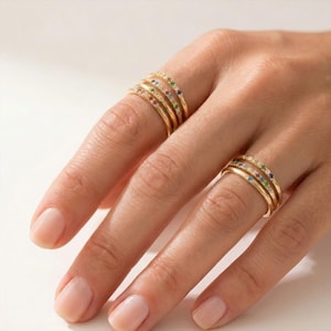 Puede incluir: Anillos dorados con piedras preciosas de colores se muestran en una mano. Los anillos son bandas finas, cada uno adornado con una fila de pequeñas gemas vibrantes en varios colores. Las piedras preciosas incluyen piedras rojas, azules, verdes y claras.