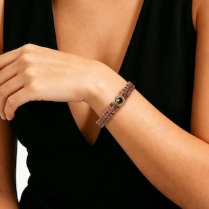 Puede incluir: Pulsera de macramé marrón con cierre dorado y piedra negra. La pulsera se lleva en la muñeca. El macramé está tejido apretado y el cierre es circular. Un accesorio de moda.