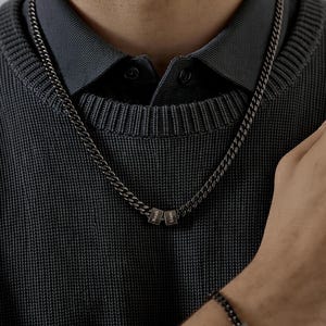 Puede incluir: Un collar de cadena gris oscuro con tres pequeños colgantes cuadrados. El collar se lleva sobre un suéter y una camisa con cuello gris oscuro. Los colgantes tienen texto grabado. Se ve una pulsera a juego.