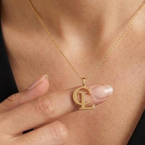 Puede incluir: Un collar de tono dorado con un colgante que presenta las letras mayúsculas C y L superpuestas. El colgante cuelga de una delicada cadena de tono dorado. El diseño consiste en una letra C grande con una letra L más pequeña en su interior.