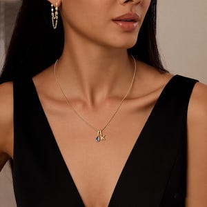 Puede incluir: Un collar de oro con un colgante con la letra "K" y una piedra preciosa azul. La imagen también muestra pendientes de oro con múltiples eslabones de cadena. La mujer lleva un vestido negro.