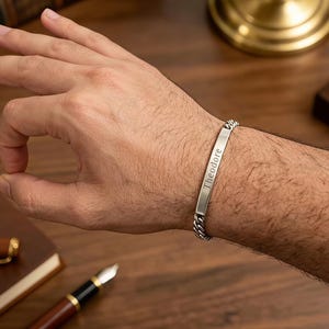 Puede incluir: Una pulsera de cadena de metal en tono plateado con una placa de identificación rectangular y plana, grabada con el nombre Theodore en una fuente oscura y clara. La pulsera consta de eslabones entrelazados y un cierre metálico.