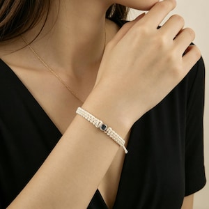 Puede incluir: Pulsera de macramé color crema con una piedra gris oscuro en el centro. La pulsera se lleva en la muñeca y también se ve un collar de oro. El fondo es una blusa negra.