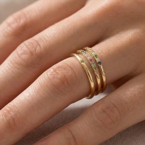 Puede incluir: Tres anillos de oro apilados en un dedo. Un anillo es liso, mientras que los otros dos están adornados con pequeñas piedras preciosas de colores. Las piedras preciosas incluyen verde, azul, rosa y amarillo. Los anillos son delicados y elegantes.
