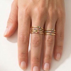 Puede incluir: Una mano que lleva varios anillos de oro. Cada anillo es delgado y está adornado con pequeñas piedras preciosas de colores. Las piedras preciosas incluyen rojo, verde, azul y blanco. Los anillos están apilados en los dedos, creando un aspecto en capas.