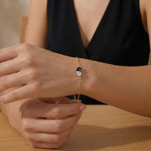 Puede incluir: Una delicada pulsera dorada con una cadena y dos piedras preciosas, una azul y otra transparente. La pulsera se lleva en la muñeca, mostrando su fino diseño. La pulsera es una pieza de joyería.