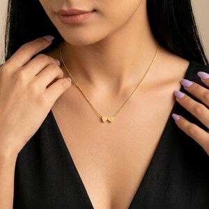 Puede incluir: Un collar dorado con una cadena delicada y dos colgantes en forma de corazón. Cada colgante tiene una letra grabada. El collar se lleva sobre una blusa negra.