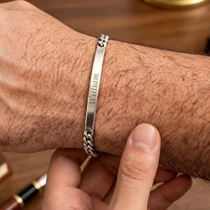 Puede incluir: Una pulsera de identificación de metal plateado con una placa central plana y rectangular grabada con el nombre William en letra cursiva. La pulsera está construida con un diseño de cadena de eslabones tipo barbada a ambos lados de la placa.