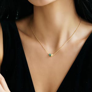 Puede incluir: Un delicado collar de oro con un pequeño colgante con detalles en verde azulado y dorado. El collar se lleva sobre una parte superior negra, resaltando el diseño de la joya. La cadena es fina y el colgante es el punto focal.