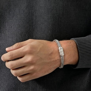 Puede incluir: Una pulsera de cadena trenzada en tono plateado con tres cuentas cilíndricas grabadas con los nombres LIAM, NOAH y OLIVER. La pulsera tiene un diseño de metal tejido texturizado y se lleva en la muñeca sobre un suéter gris oscuro.
