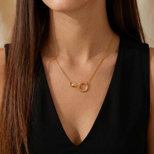 Puede incluir: Un collar dorado con una cadena delicada y un colgante. El colgante presenta un diseño circular y un elemento más pequeño adjunto con el nombre "Isabela" grabado. El collar se lleva sobre una blusa negra.
