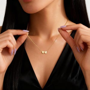 Puede incluir: Un delicado collar de oro con dos colgantes en forma de corazón. Cada colgante tiene una inicial. El collar se sostiene sobre un fondo negro. La cadena es fina y los colgantes son pequeños.