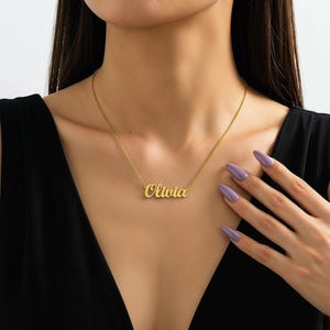 Puede incluir: Un collar dorado con el nombre "Olivia" en cursiva. El collar se lleva alrededor del cuello. La persona lleva una blusa negra y tiene el pelo largo y oscuro. La mano de la persona es visible, con uñas largas y moradas.