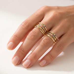 Puede incluir: Anillos dorados con piedras preciosas de colores. Los anillos están apilados en una mano, mostrando una variedad de colores, incluyendo rojo, azul, verde y amarillo. Los anillos tienen un diseño delicado y minimalista.