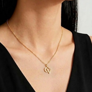 Puede incluir: Un collar de tono dorado con una cadena de eslabones trenzados y un colgante que muestra las letras C y V entrelazadas. El colgante está pulido y cuelga en el centro de la cadena, que descansa sobre el cuello con un diseño clásico y sencillo.