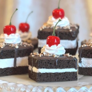 May include: Several chocolate cake slices with white cream filling and a red cherry on top are arranged on a white, decorative cake stand. The cakes have a chocolate glaze and are sprinkled with nuts.
