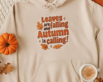 Puede incluir: Sudadera con capucha beige con bolsillo delantero y cordón. La parte delantera presenta el texto "Leaves are falling and Autumn is calling!" en naranja y marrón. Se ven una pequeña calabaza y una taza de café.