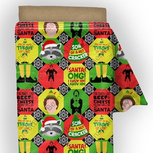 May include: A festive Christmas-themed blanket with a repeating pattern of red, green, and yellow squares. The design includes images of a raccoon in a Santa hat, elves, and text phrases like "Santa OMG!" and "You smell like beef & cheese."