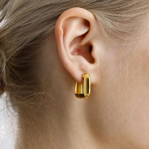 Op de afbeelding: Close-up van een oor met een paar goudkleurige rechthoekige oorringen. De oorbellen hebben een gepolijste afwerking en zijn een modern sieradenontwerp.