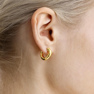 Könnte beinhalten: Nahaufnahme eines goldfarbenen Creolen-Ohrrings, der an einem Ohr getragen wird. Der Ohrring hat ein dickes, abgerundetes Design und ein glänzendes Finish. Das Ohr ist an der Haut einer Person befestigt, und der Hintergrund ist verschwommen.