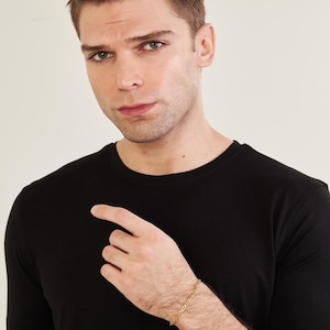 May include: A gold chain bracelet is worn on a person's wrist. The bracelet features a link design. The person is wearing a black t-shirt. The background is a plain, light colour.