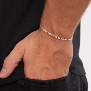 May include: A silver chain bracelet with a curb link design. The bracelet is worn on a wrist, with the hand in a pocket. The bracelet is a simple, elegant piece of jewelry.