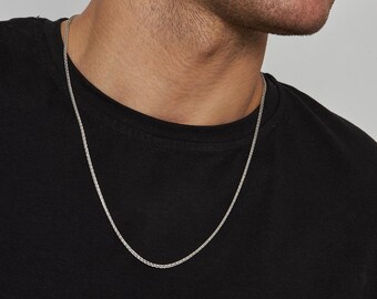 Könnte beinhalten: Eine silberne Halskette mit gewebtem Design. Die Halskette wird vor einem schwarzen T-Shirt präsentiert, was ihren metallischen Glanz hervorhebt. Ein klassisches Accessoire.