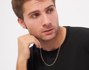 Könnte beinhalten: Eine goldfarbene Halskette. Die Halskette ist ein schlichtes, klassisches Design, wahrscheinlich aus Gold oder vergoldetem Metall. Die Kette wird um den Hals getragen, auf einem schwarzen T-Shirt liegend.