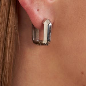 May include: A close-up of a silver square hoop earring. The earring is a simple, geometric design with a polished finish. The earring is worn in an ear, with the skin and hair visible in the background.