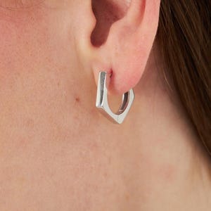 May include: A silver-toned hoop earring with a distinct hexagonal shape. The earring features a polished metallic finish and a hinged closure mechanism, designed to sit securely through the earlobe with a clean, geometric aesthetic.