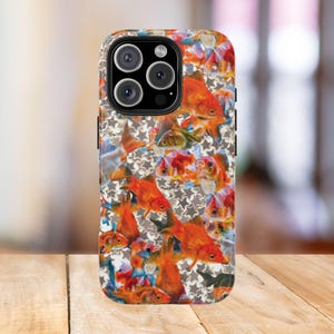 Puede incluir: Una funda de teléfono con un vibrante diseño de peces dorados naranjas sobre un fondo de formas de estrellas grises. La funda tiene un módulo de cámara negro con tres lentes y un flash. La funda está sobre una superficie de madera.