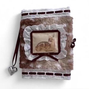 May include: A rectangular journal covered in brown and white faux fur, featuring a central rectangular patch with a fawn illustration. The cover is decorated with white lace trim, dark brown ribbon accents, and a silver heart-shaped charm.