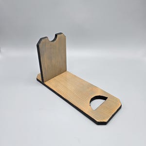 May include: A wooden phone stand with a natural wood finish and a dark gray trim. The stand has a rectangular base with a curved cutout and a vertical back with a notch. The stand is designed to hold a phone.