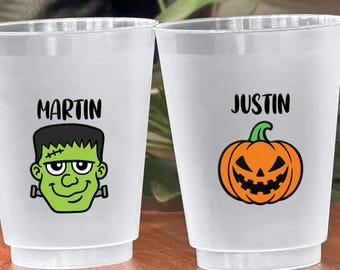May include: Two clear plastic cups with Halloween-themed designs. One cup features a green Frankenstein's monster graphic with the name "MARTIN" above it. The other cup has an orange jack-o'-lantern graphic with the name "JUSTIN".