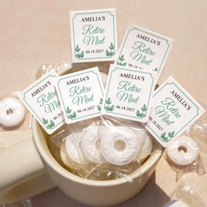 May include: A ceramic mug filled with white mint candies, some in individual clear wrappers. The candies are topped with small square labels that read "AMELIA'S Retire Mint 06.18.2027" in a decorative font with green accents.