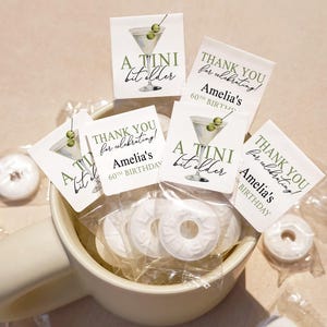May include: A white ceramic mug filled with individually wrapped white mint candies. Several small square tags with a martini illustration and text that reads "A Tini bit older" and "Thank you for celebrating! Amelia's 60th Birthday" are scattered around the mug.