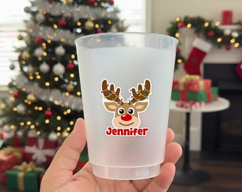 May include: A clear plastic cup with a reindeer graphic and the name "Jennifer" in red. The reindeer has brown antlers decorated with Christmas lights and a red nose. The cup is held in front of a Christmas tree.