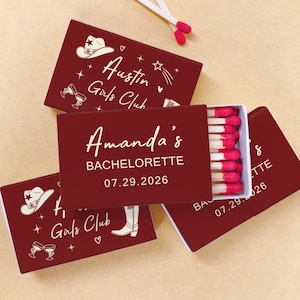 May include: A collection of burgundy matchboxes with white text and illustrations. One box is open, revealing red-tipped matches. The text reads "Austin Girls Club" and "Amanda's BACHELORETTE 07.29.2026". The design includes a cowboy hat, boots, and cocktail glasses.