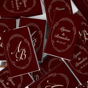 May include: A collection of rectangular matchboxes in a deep burgundy color. Each box features a gold-colored ornate oval frame containing either the names Alyssa & Brandon with the date 06.25.27 or the intertwined initials AB in a script font.