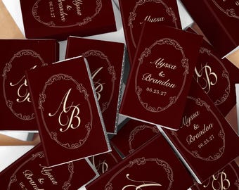 Puede incluir: Una colección de cajas de cerillas rectangulares en color burdeos intenso. Cada caja presenta un marco ovalado ornamentado de color dorado que contiene los nombres Alyssa & Brandon con la fecha 06.25.27 o las iniciales entrelazadas AB.