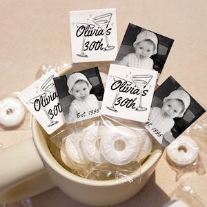 May include: A light-colored mug filled with individually wrapped white ring-shaped candies. Each candy features a square tag with black text reading Olivia's 30th, Est. 1996, and a black and white photograph of a young child wearing a knit hat.