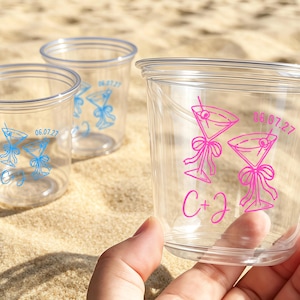 May include: Clear plastic cups with a printed cocktail design in pink and blue. The cups have the date "06.07.27" and initials "C + J" printed on them. The cups are sitting on a sandy surface.
