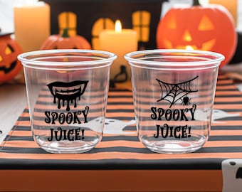 May include: Two clear plastic cups with black graphics and text that read "Spooky Juice!". One cup features vampire fangs, the other a spider web with a spider. The cups are on a Halloween-themed table runner.