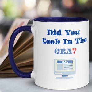 Peut inclure: Mug en céramique blanche avec une anse et un intérieur bleu marine. Le mug affiche le texte "Did You Look In The CBA ?" en bleu, avec un graphique d'ordinateur et le texte "ITAS" en dessous. Un livre est en arrière-plan.