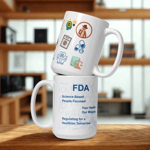 May include: Two white ceramic mugs stacked on a wooden surface. The top mug features various medical-related icons and a cartoon illustration. The bottom mug displays the FDA logo and the text "Science Based People Focused" and "Regulating for a Healthier Tomorrow."